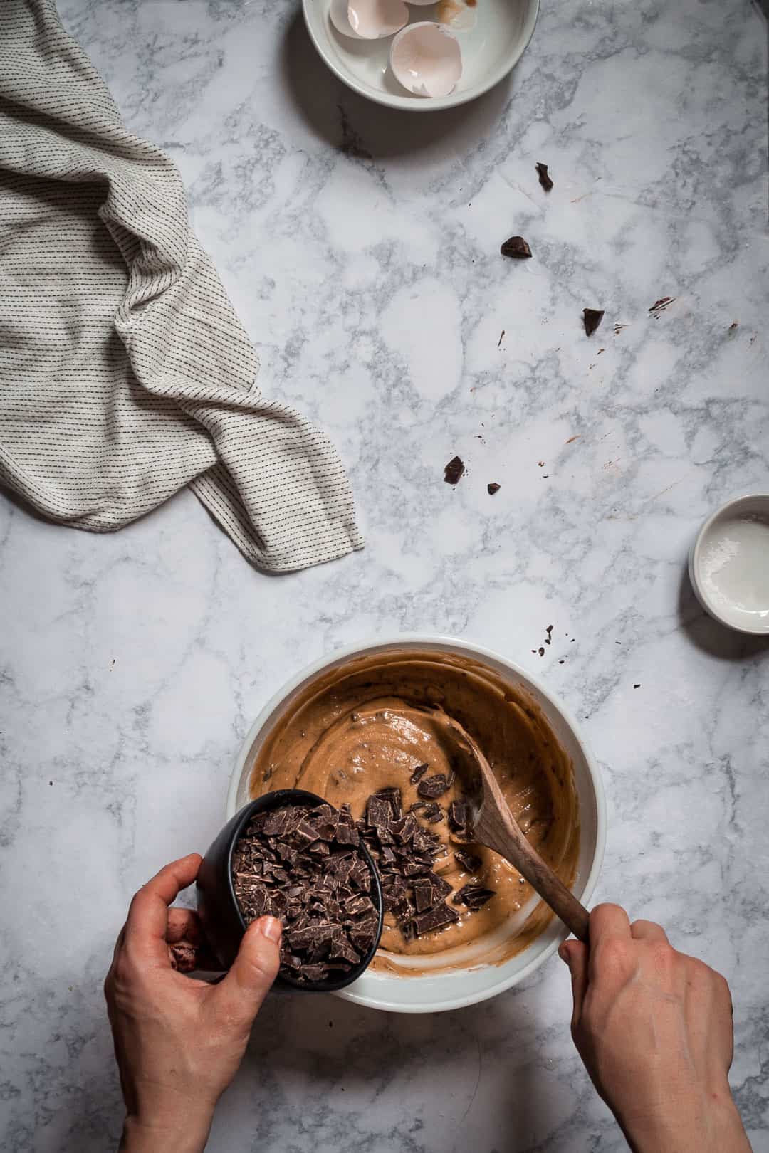 process shot of hands pouring dark chocolate chunks into a bowl of butterscotch bar cookie batter