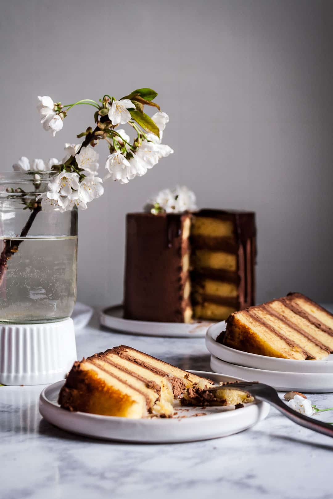 marzipan cake with chocolate buttercream and chocolate ganache drip on a white platter and marble surface, with two pieces cut and plated on white plates next to a vase of white cherry blossoms