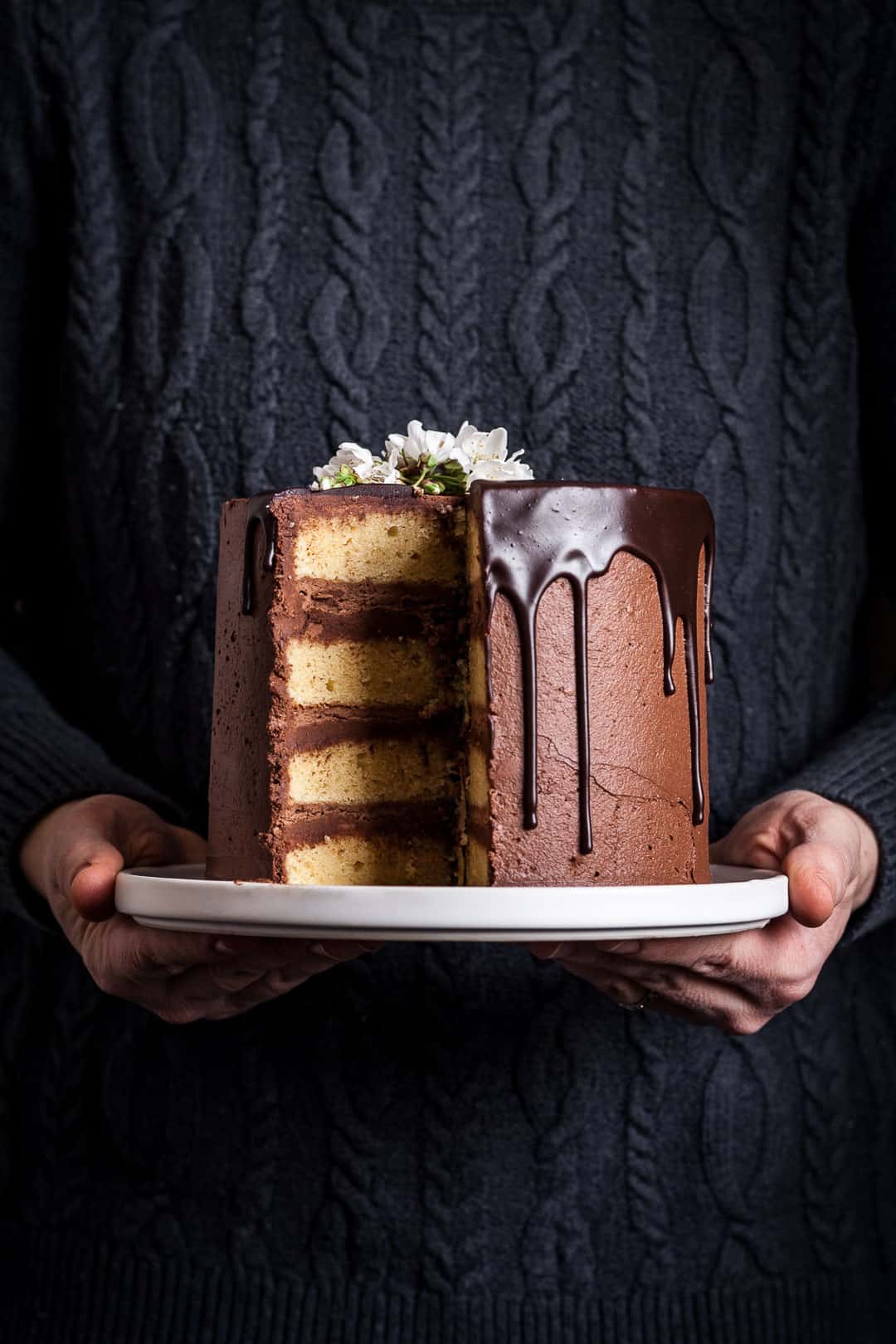 closeup of yellow marzipan cake with dark chocolate buttercream with a slice missing, on a white platter being held by a person wearing a navy blue cable knit sweater