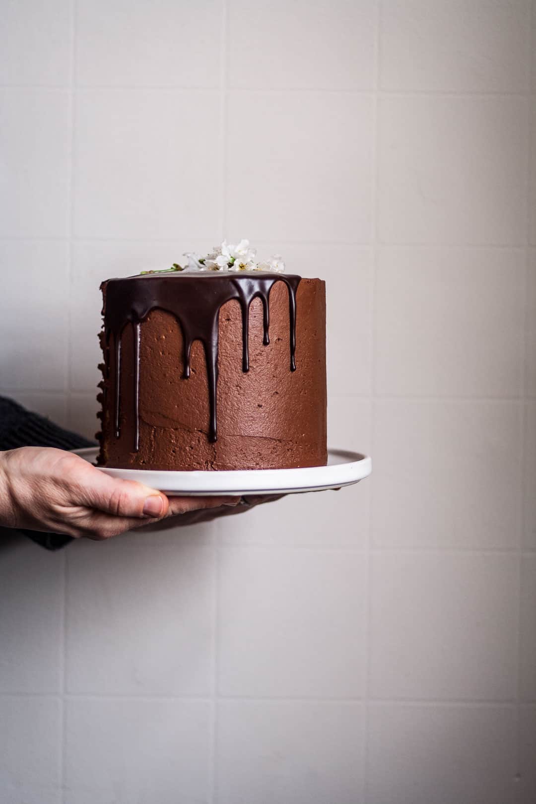 two hands holding marzipan cake with chocolate buttercream and chocolate ganache drip on a white platter against a white wall