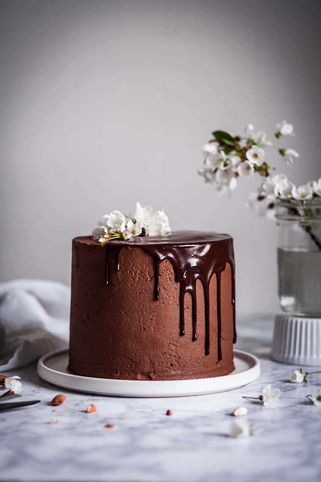 marzipan cake with chocolate buttercream and chocolate ganache drip on a white platter and marble surface, next to a vase of white cherry blossoms