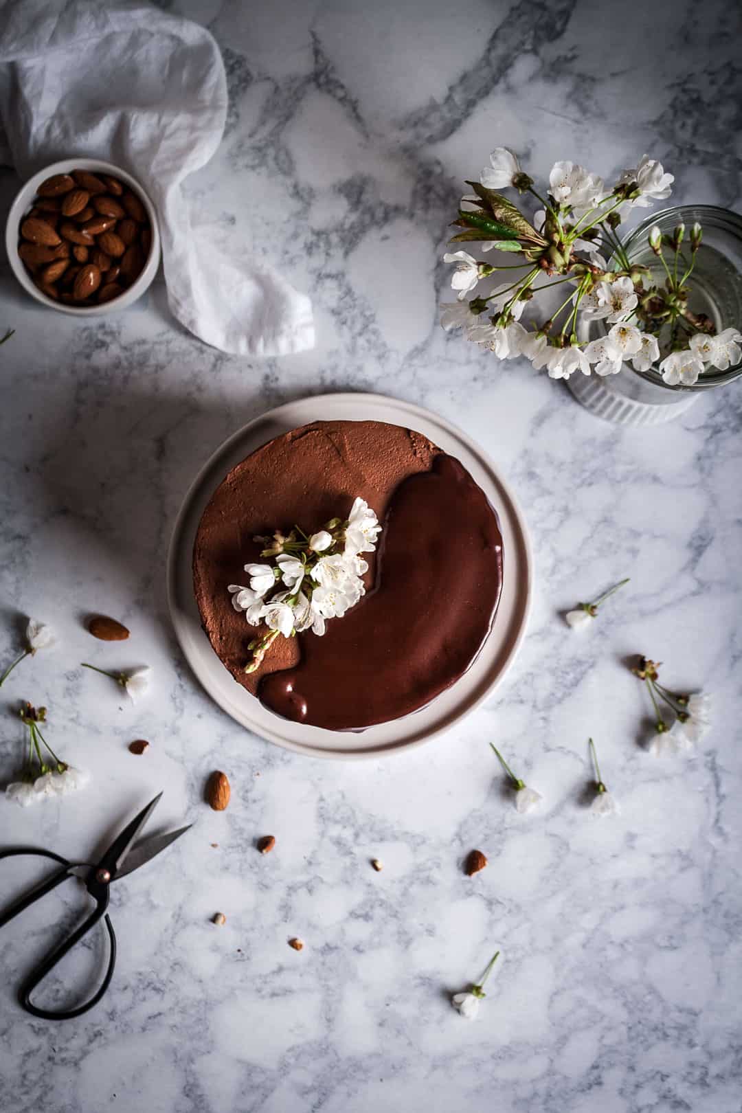 Top view of marzipan layer cake with dark chocolate buttercream and chocolate ganache drip, on a marble surface with white cherry blossoms and a pair of garden shears