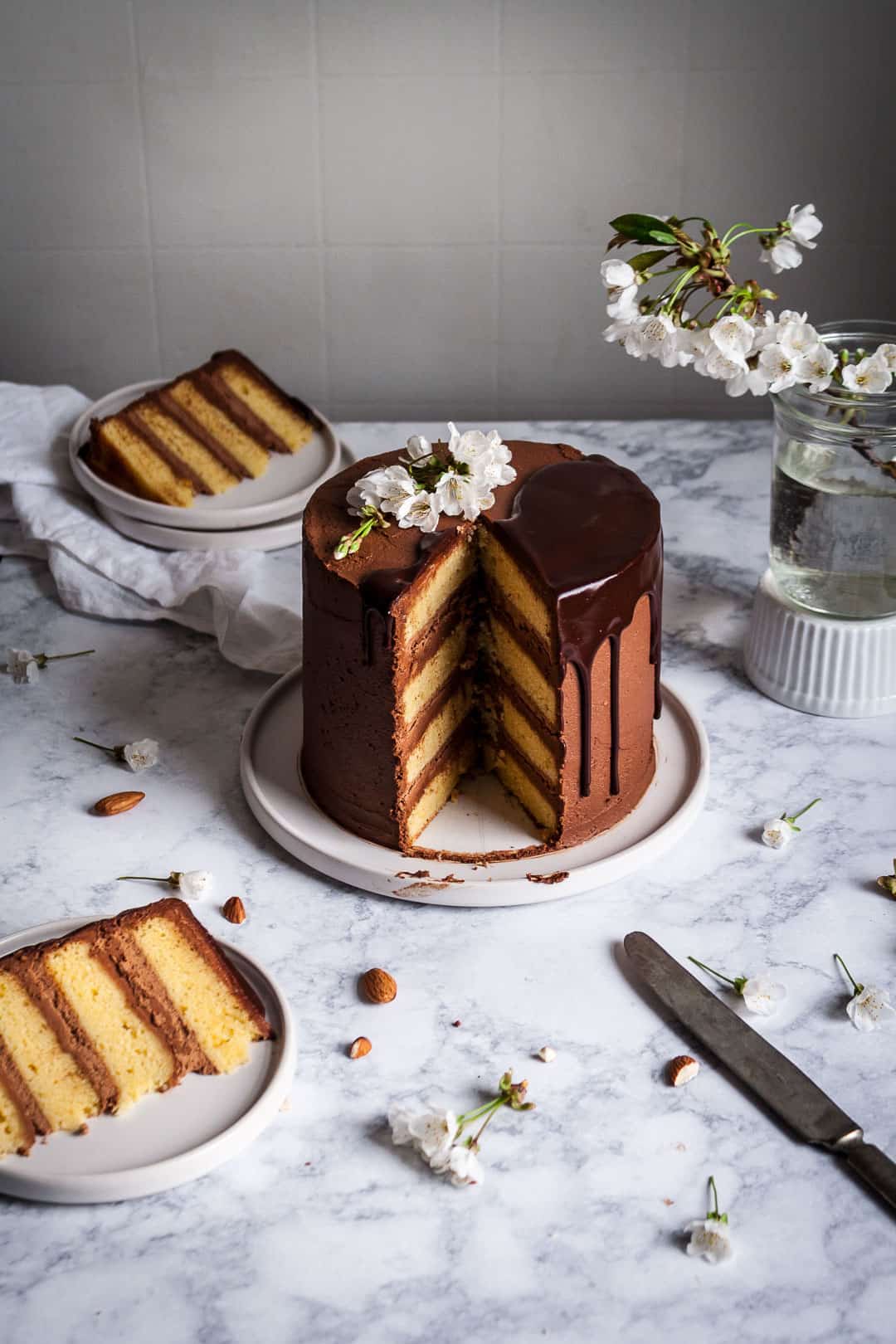 side view of marzipan cake with dark chocolate buttercream with two slices cut out and plated next to the cake on a marble surface