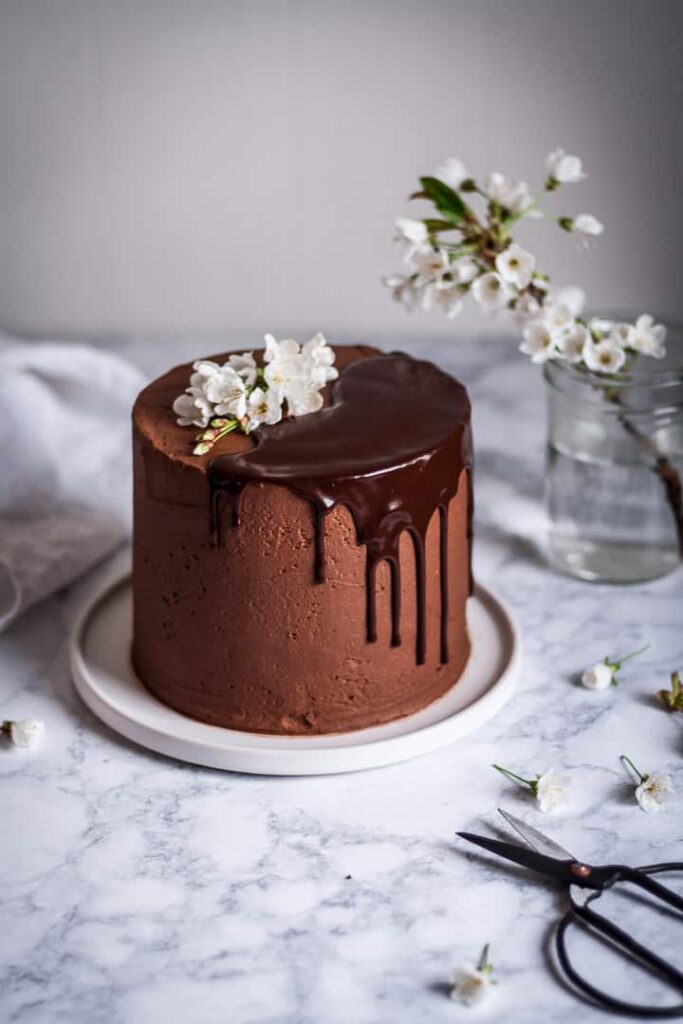 marzipan cake with chocolate buttercream and chocolate ganache drip on a white platter and marble surface, next to a vase of white cherry blossoms