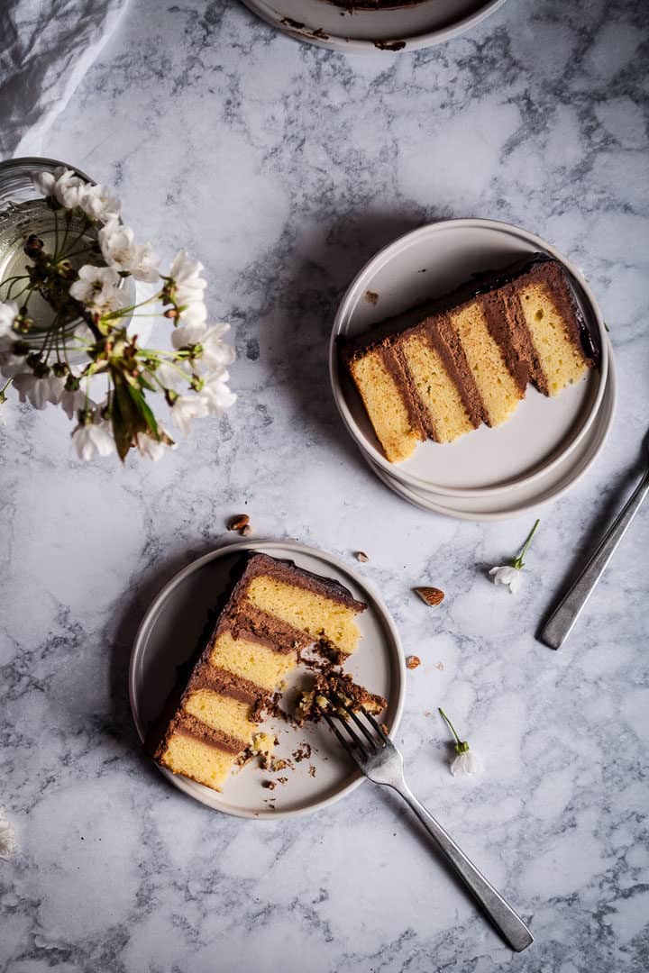 top view of two slices of marzipan cake with dark chocolate buttercream, on white plates with forks on a marble countertop