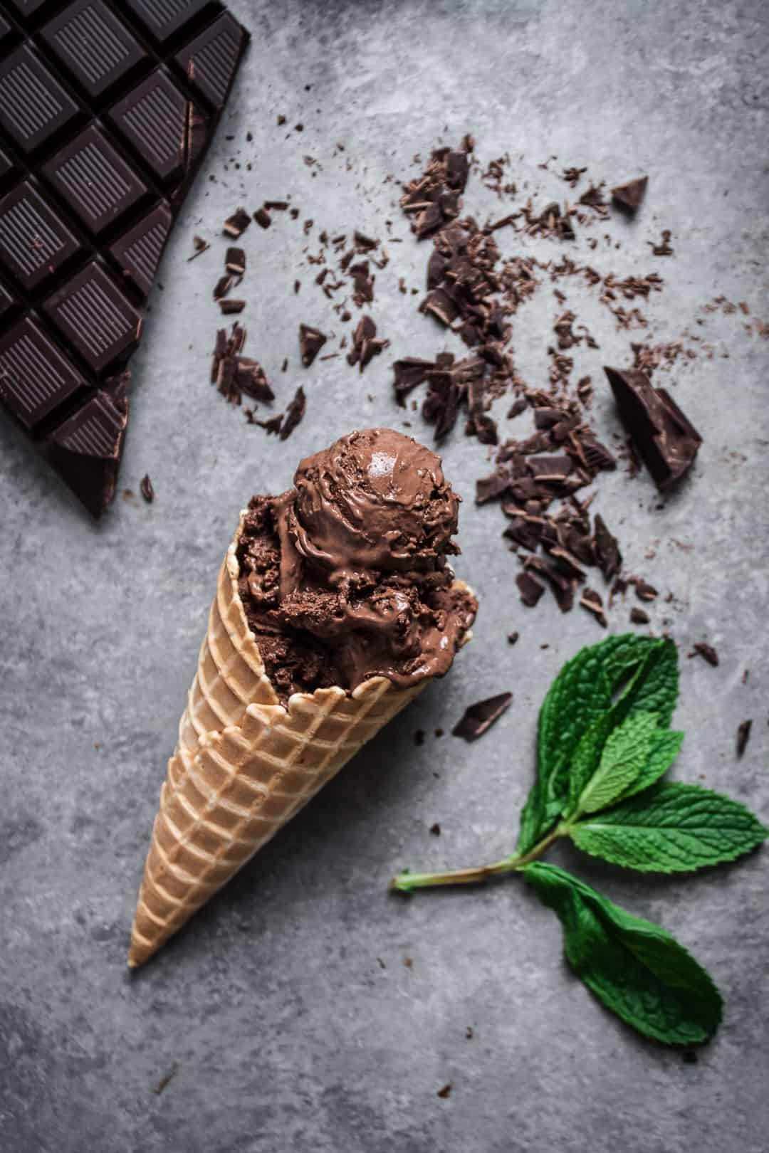 top view of mint mocha chocolate chip ice cream cone lying on grey background with mint leaves and chocolate bar nearby