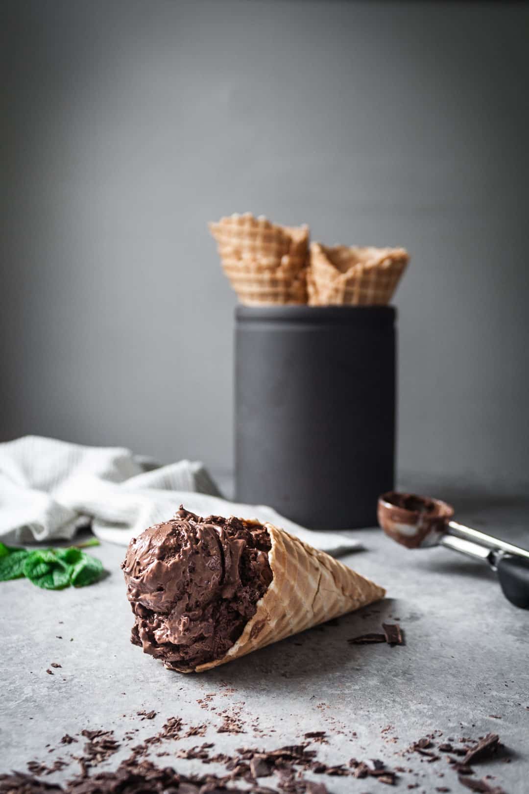 mint mocha chocolate chip ice cream in a waffle cone lying in foreground with extra waffle cones stacked in black ceramic container in background