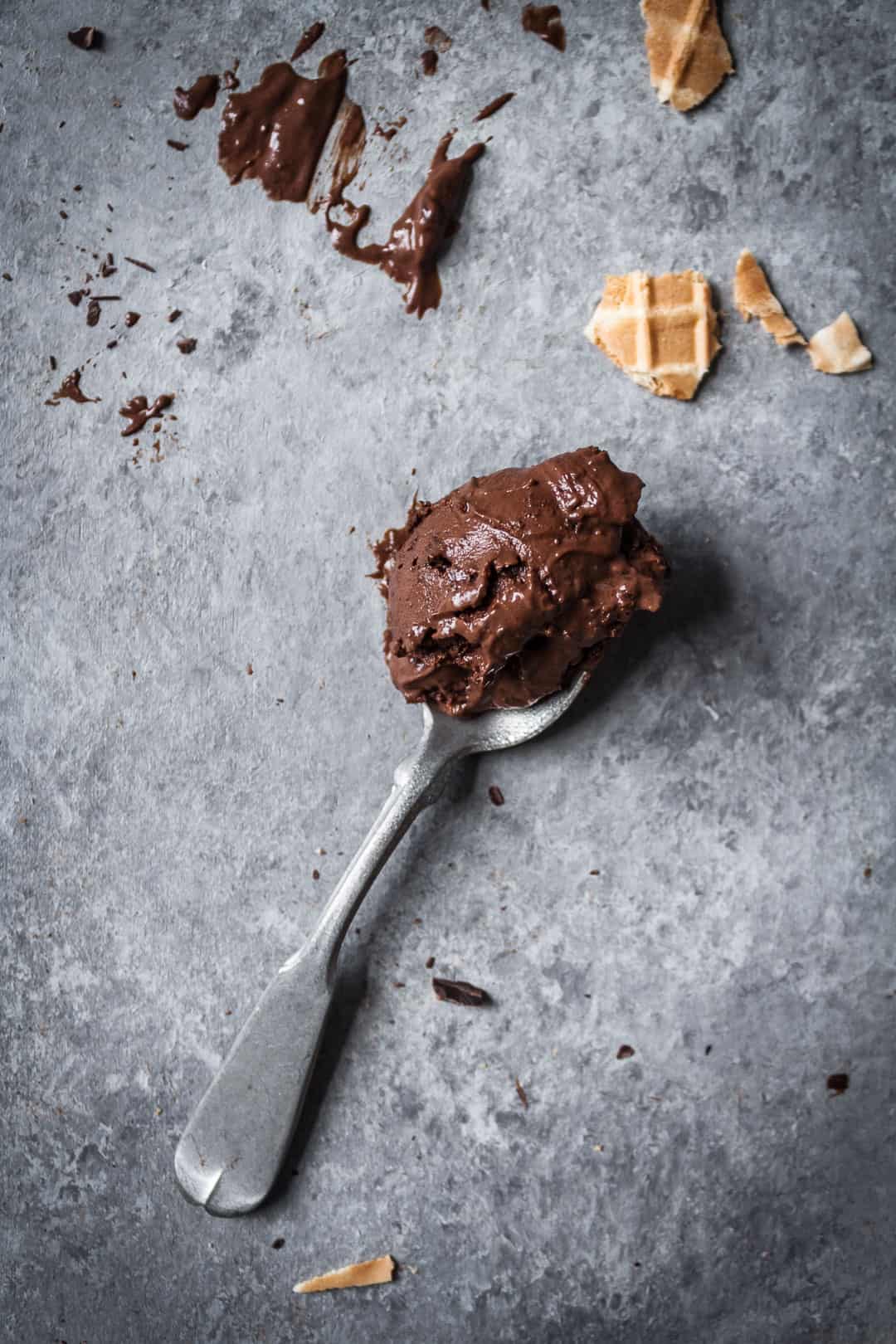 top view of a silver spoon scooped full of a bite of mint mocha chocolate chip ice cream on a grey background with chocolate pieces and broken bits of waffle cone nearby