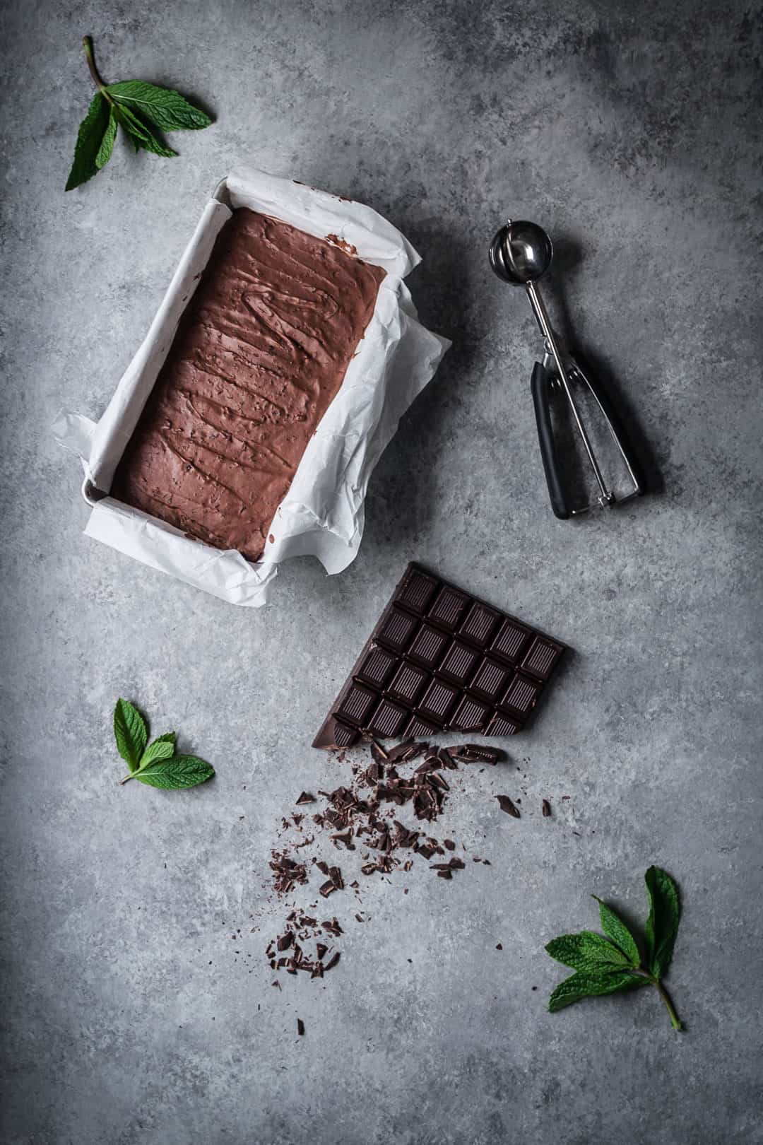 top view of mint mocha chocolate chip ice cream in container on grey background with ice cream scoop, mint leaves and chocolate bar nearby