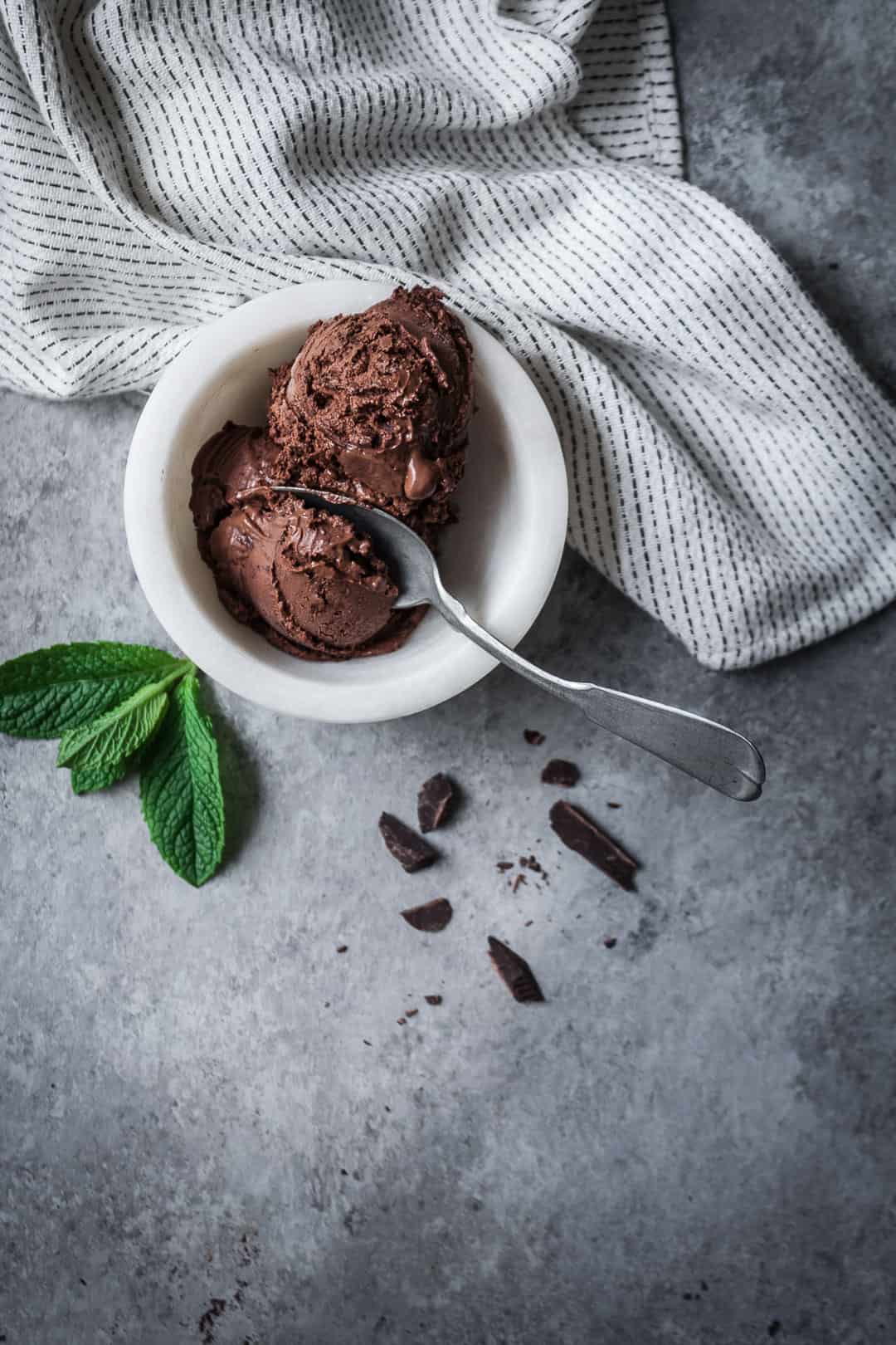 top view of white bowl of mint mocha chocolate chip ice cream with silver spoon, white kitchen towel and mint leaves on a grey background