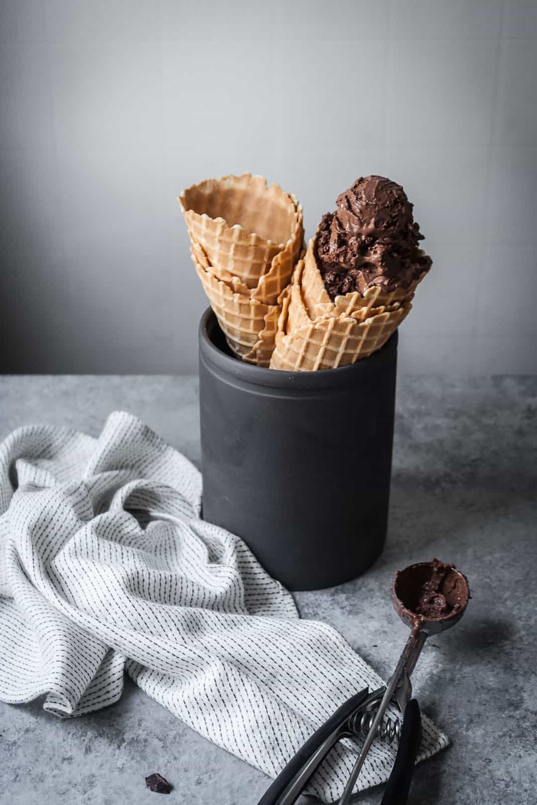front view of waffle cones in black ceramic container with white kitchen cloth and ice cream scoop nearby