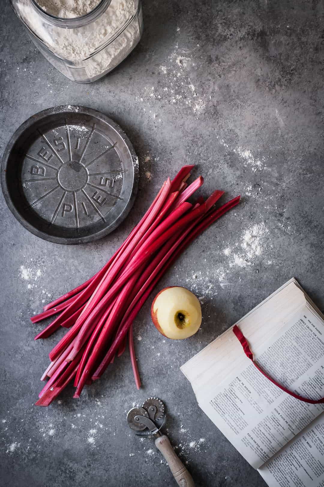 making a rhubarb apple pie with ginger and lemongrass: top view of rhubarb stalks and peeled apple on grey surface with opened cookbook, metal pie tin and flour container nearby