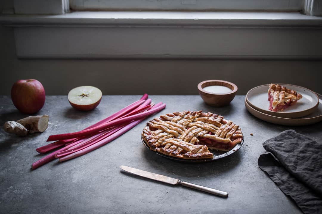 backlit image of baked rhubarb apple pie with ginger and lemongrass next to a window with a slice cut out on a plate nearby along with the pie ingredients