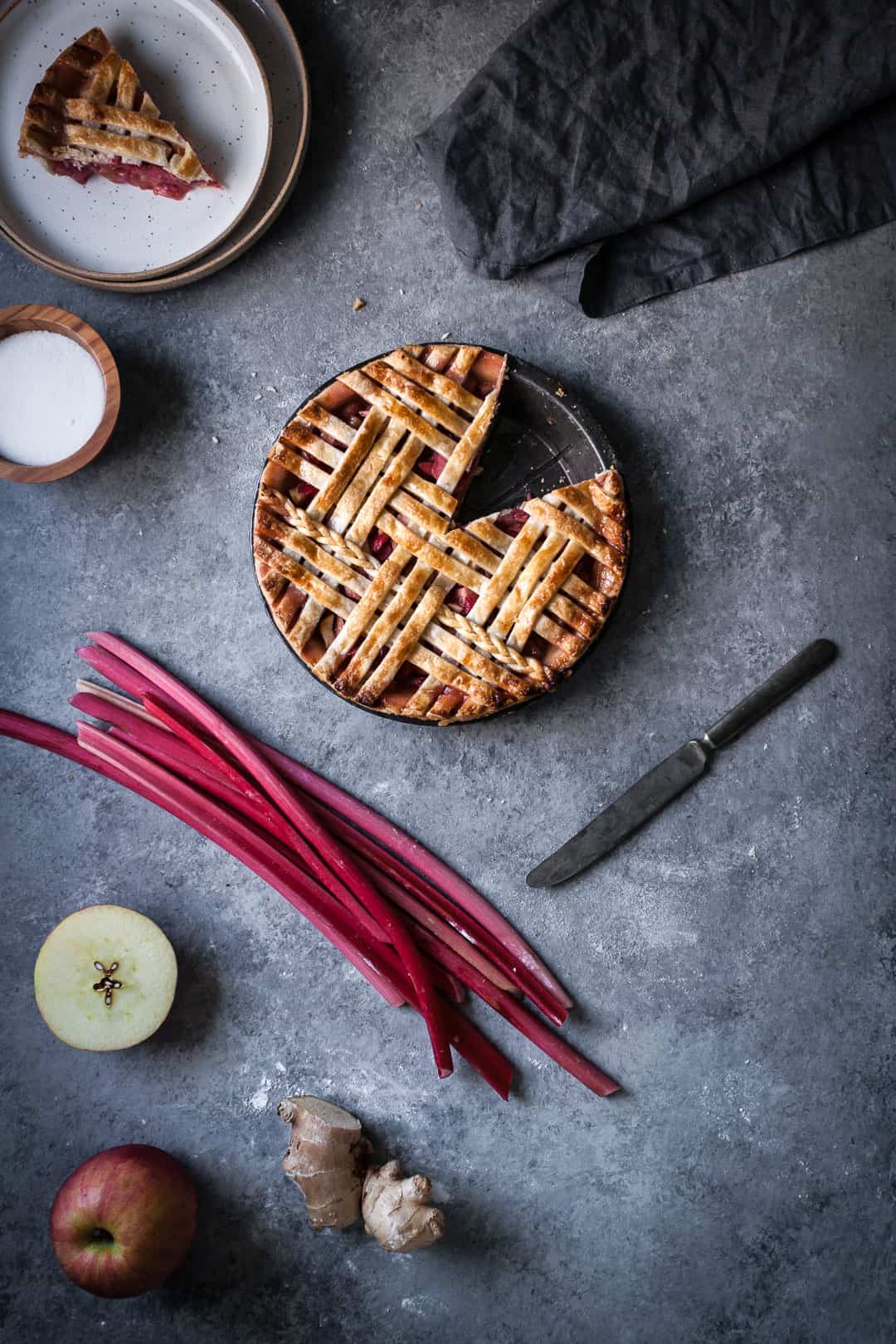 baked rhubarb apple pie with ginger and lemongrass on a grey surface with a piece cut out and the pie ingredients surrounding it