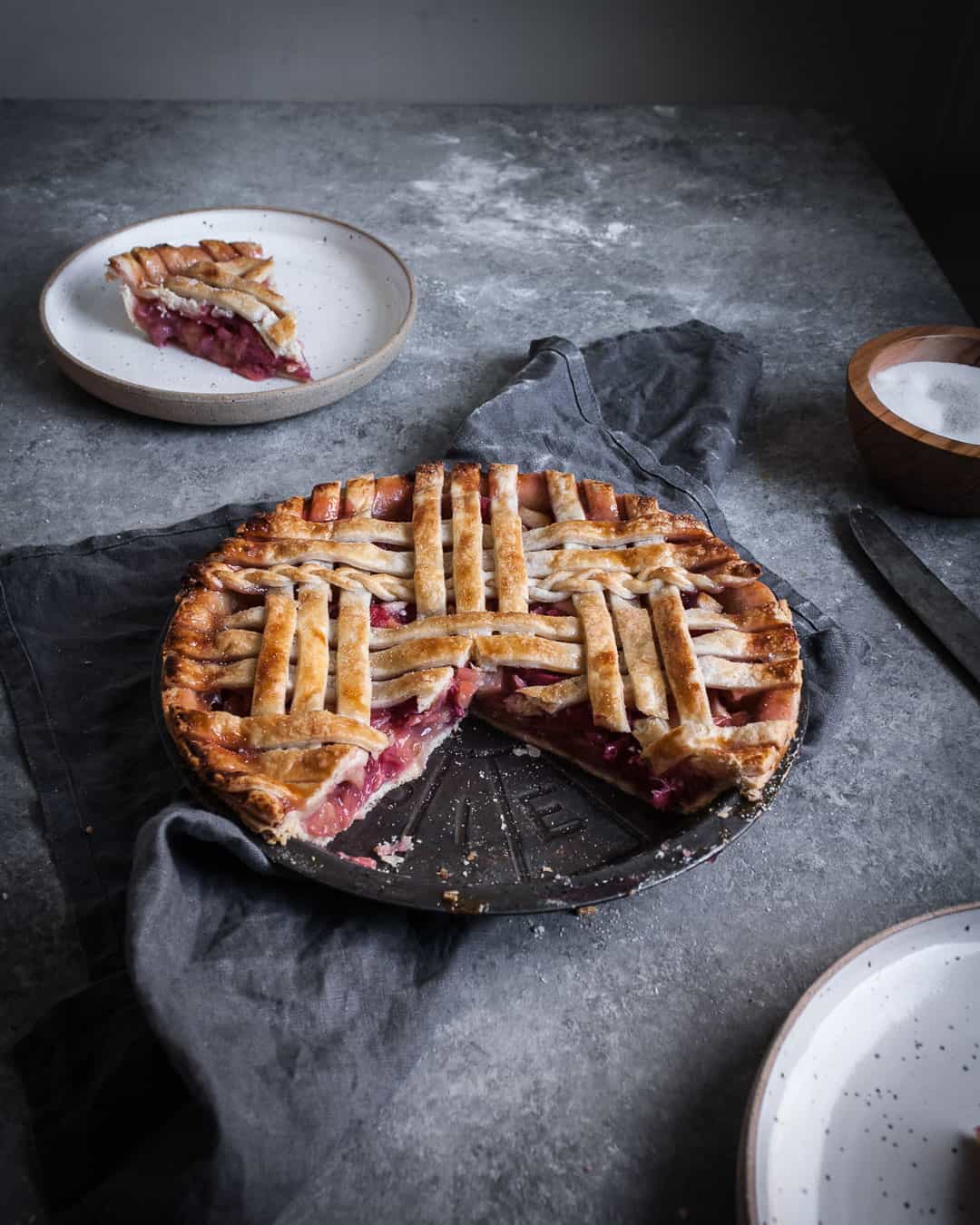 angled shot of rhubarb apple pie with ginger and lemongrass on a grey surface with a piece cut out on a plate and a grey linen towel underneath the pie tin