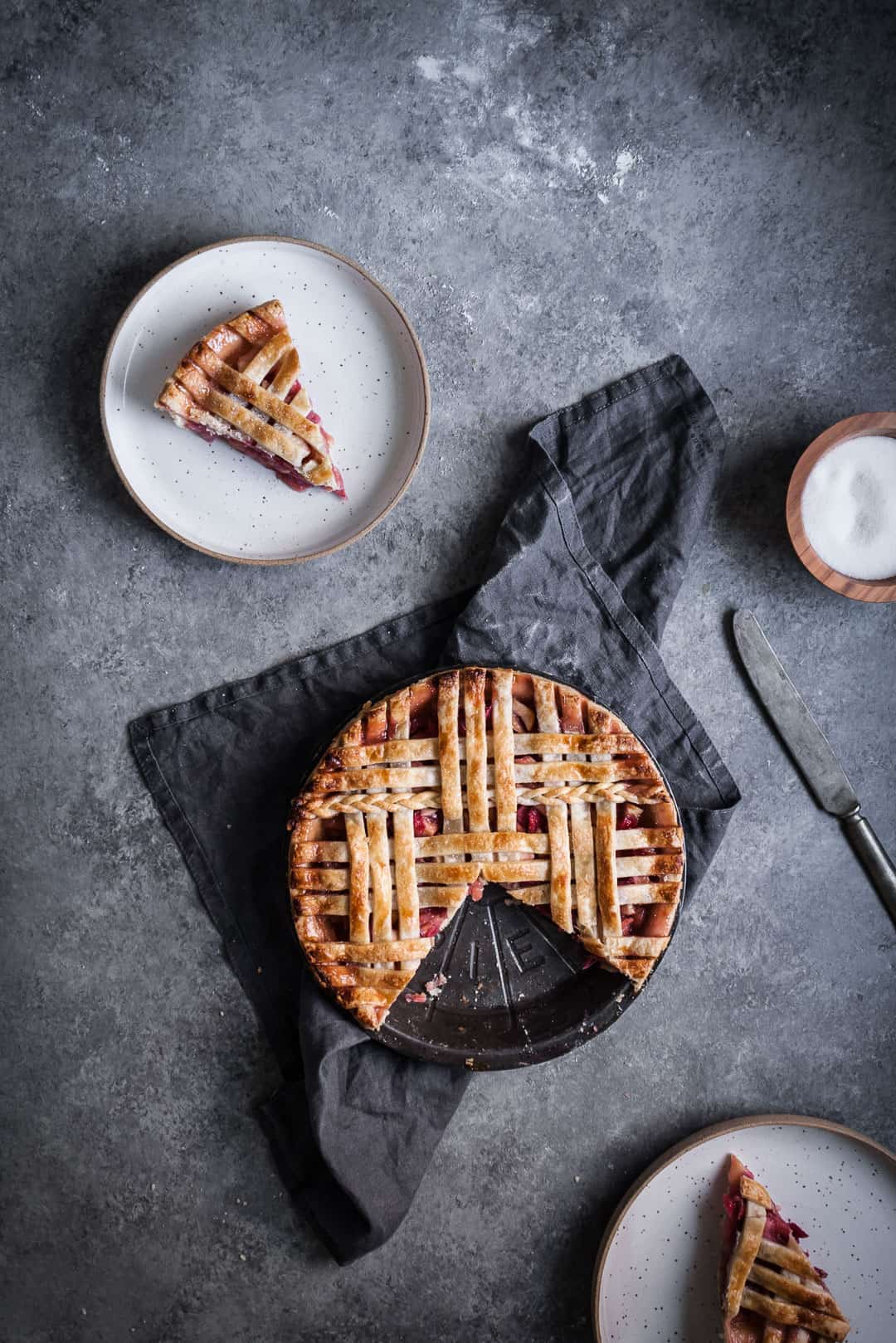 top view of baked rhubarb apple pie with ginger and lemongrass with two pieces cut out and placed on plates nearby, with a grey linen towel under the pie tin