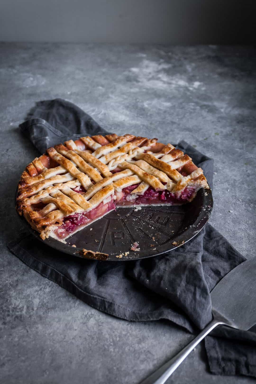 angled shot of rhubarb apple pie with ginger and lemongrass on a grey surface with a piece cut out on a plate and a grey linen towel underneath the pie tin