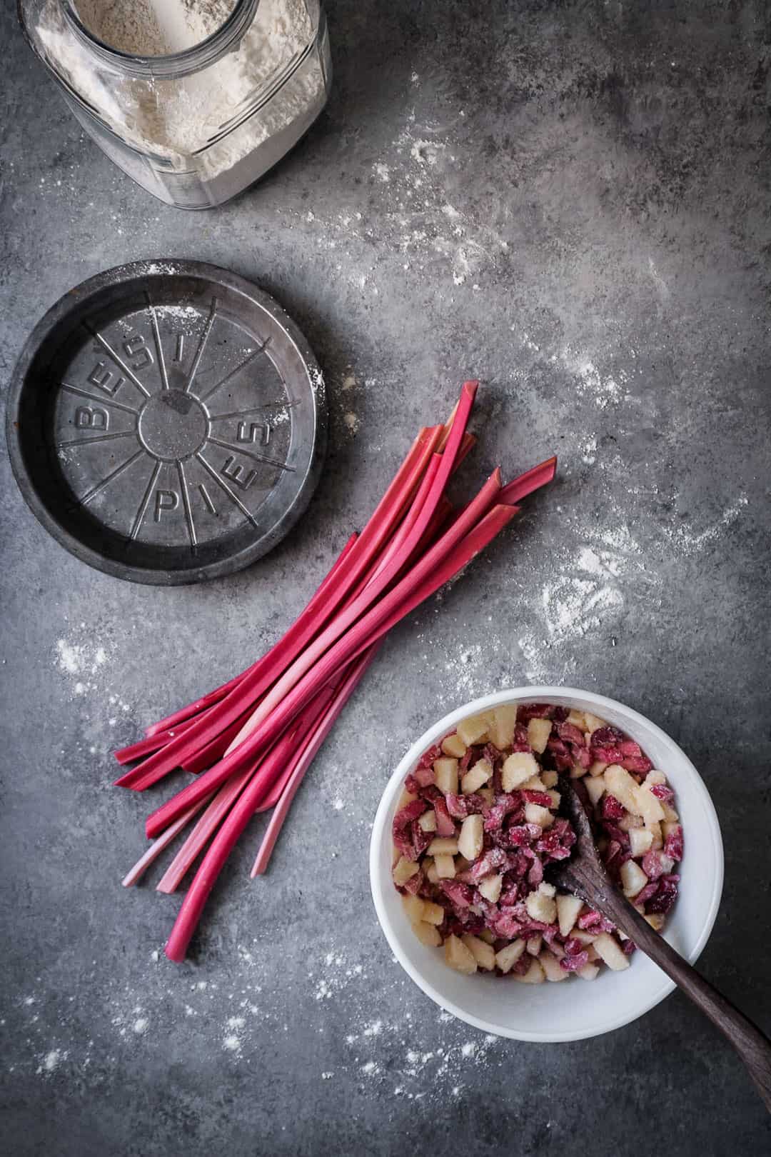 making a rhubarb apple pie with ginger and lemongrass: top view of rhubarb stalks on grey surface with metal pie tin and container of flour nearby along with white bowl filled with cut up apples and rhubarb