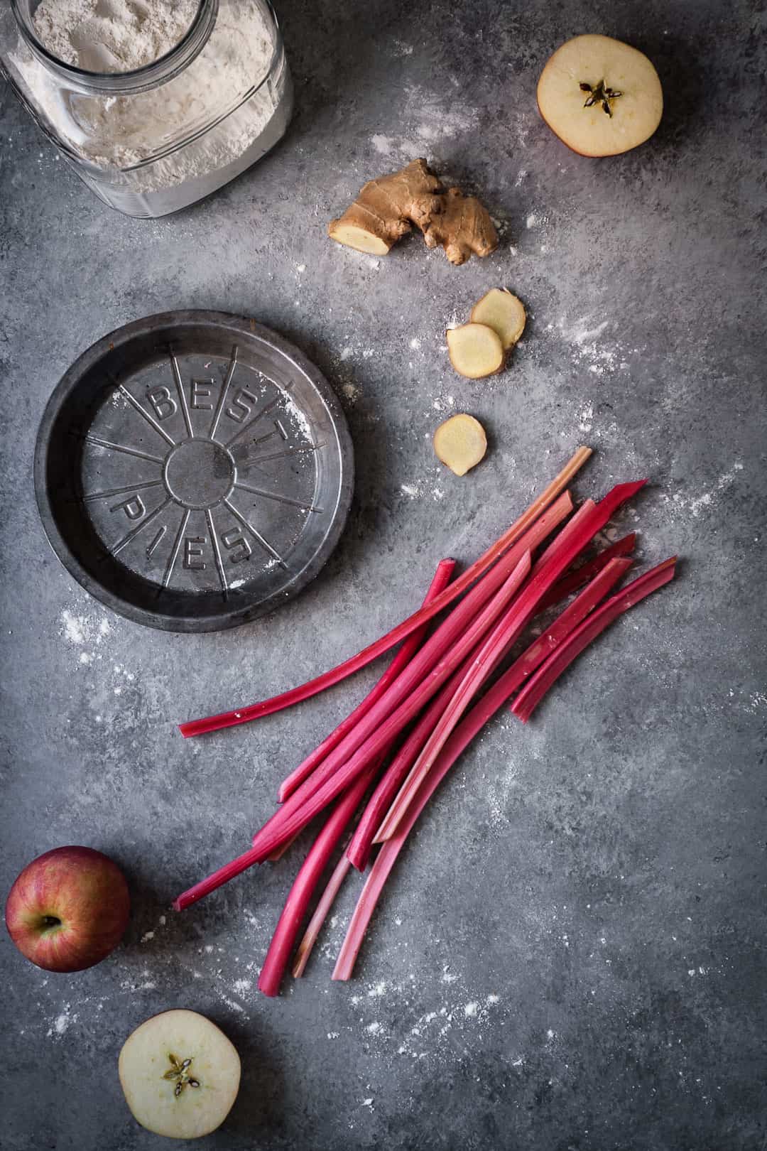 grey surface with metal pie tin and a glass jar of flour along with apples, rhubarb and ginger for rhubarb apple pie with ginger and lemongrass