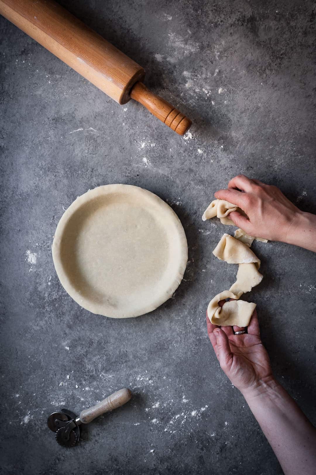 top view of hands trimming pie dough from pie tin with rolling pin lying nearby while making a rhubarb apple pie with ginger and lemongrass