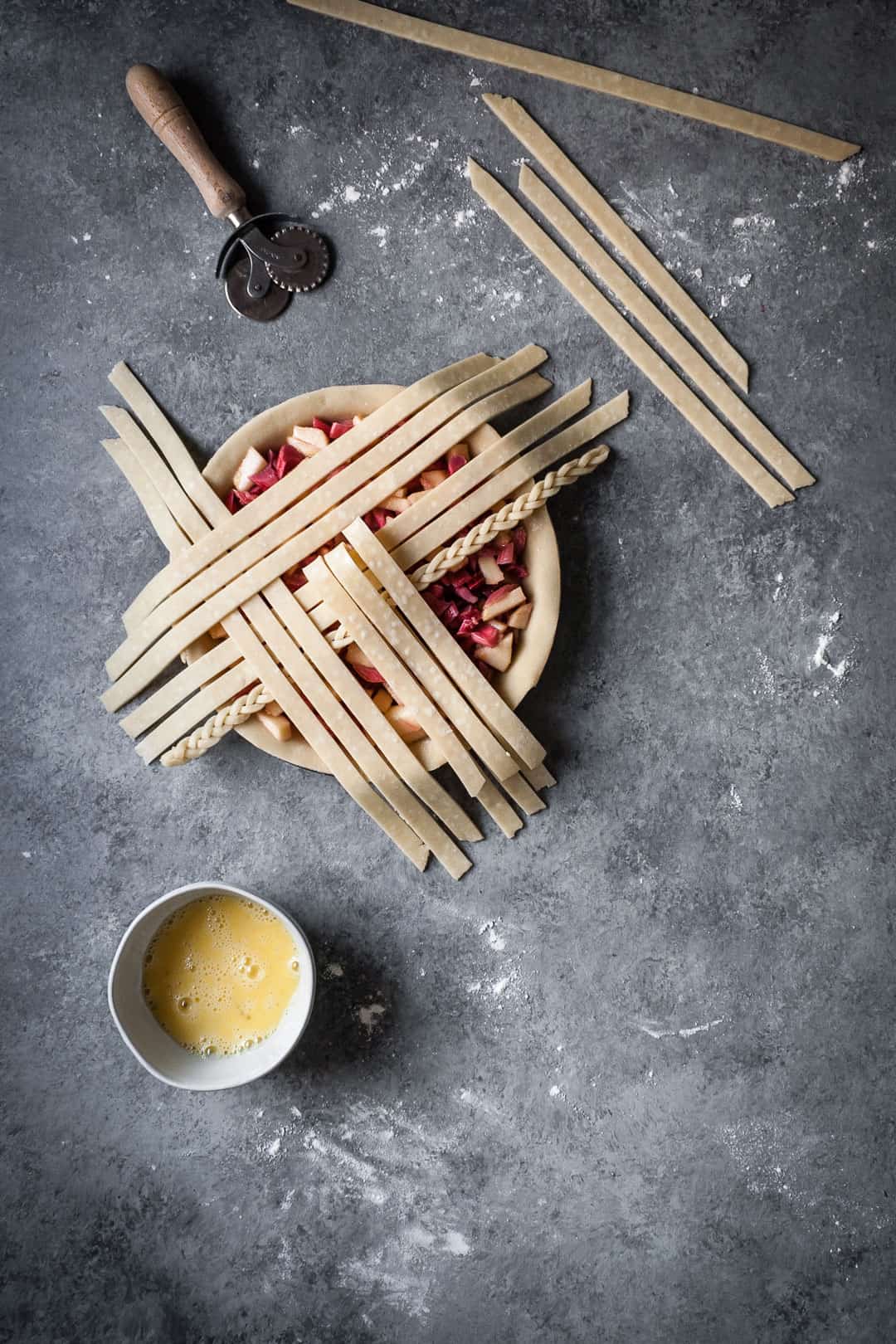 process of making a rhubarb apple pie with ginger and lemongrass - pie crust with apple rhubarb filling and lattice top in process with egg wash and dough cutter nearby