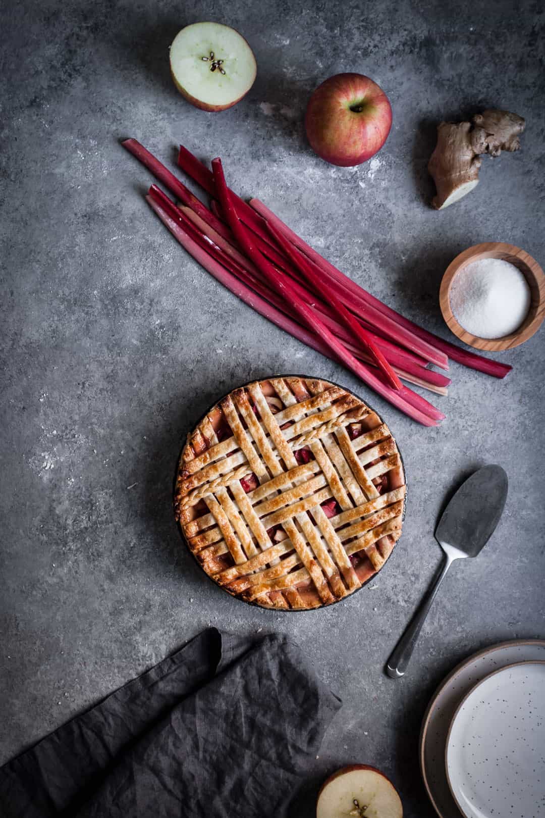 grey surface with wide view of pie making ingredients for rhubarb apple pie with ginger and lemongrass including apples rhubarb and ginger along with pie server and empty white plates