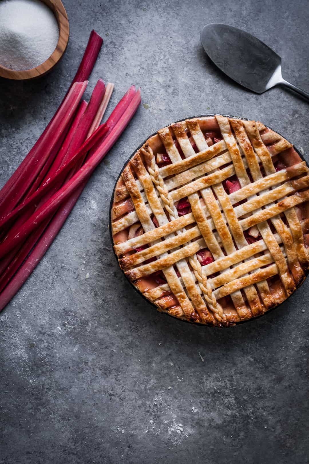 close up on grey background of baked rhubarb apple pie with ginger and lemongrass rhubarb stalks and pie server nearby