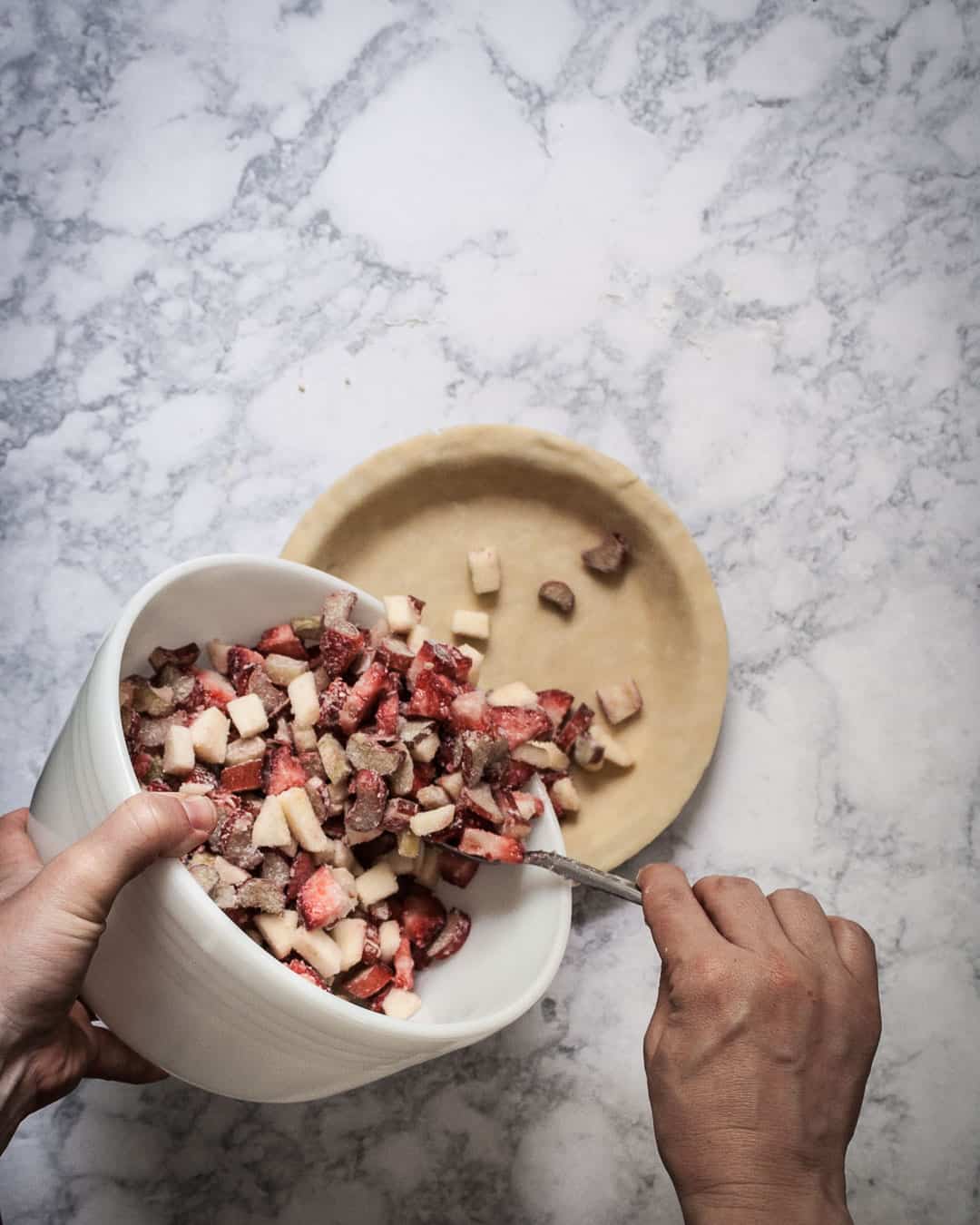 Process shot showing hands pouring a bowl of apple rhubarb pie filling into a pie crust.