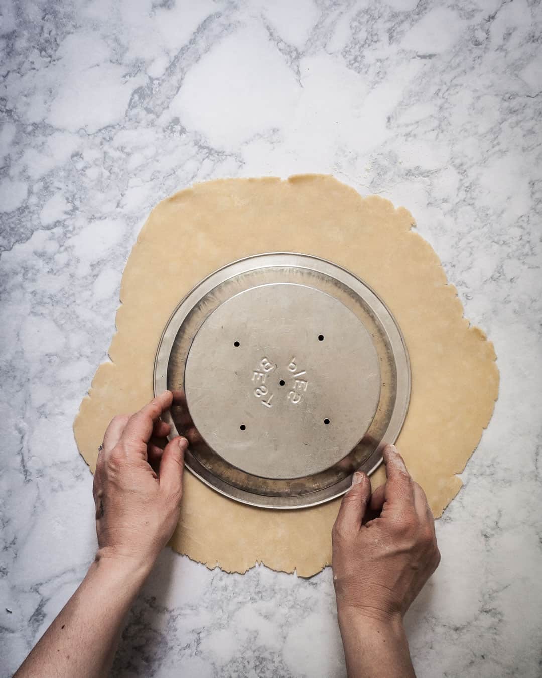 Pie crust with two hands placing a pie tin over the top to check the size of the crust.