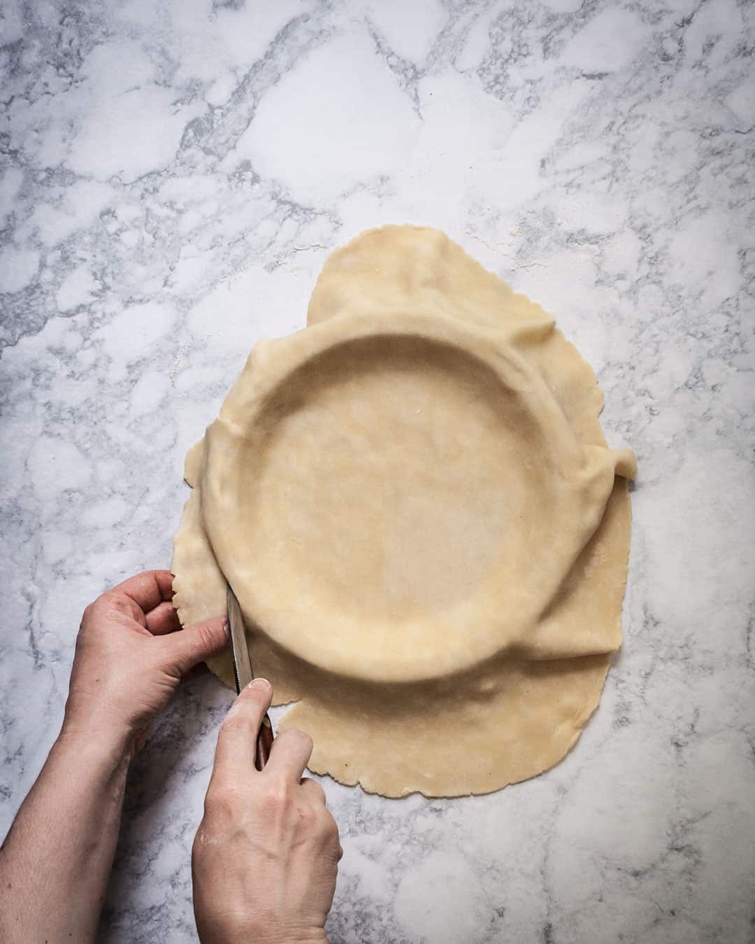 Hands trimming excess pie dough around a pie pan.