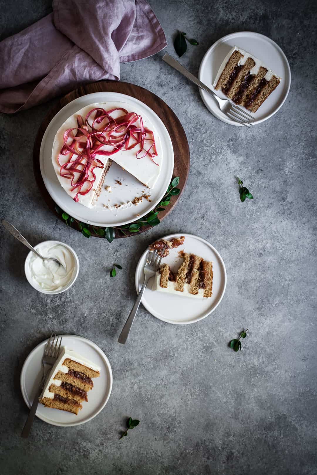 Top view table scene of brown sugar cake with rhubarb compote and creme fraiche buttercream with three slices on plates and a pot of creme fraiche