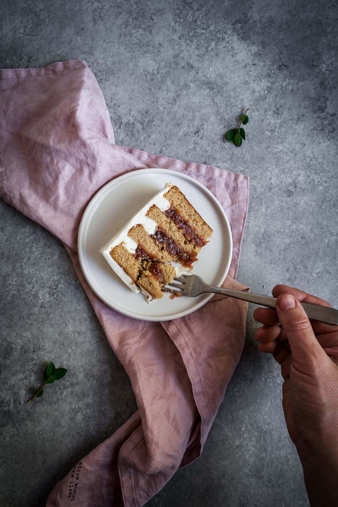 Hand holding a fork and cutting into a slice of brown sugar cake with rhubarb compote and creme fraiche buttercream on a pink linen napkin and a grey background