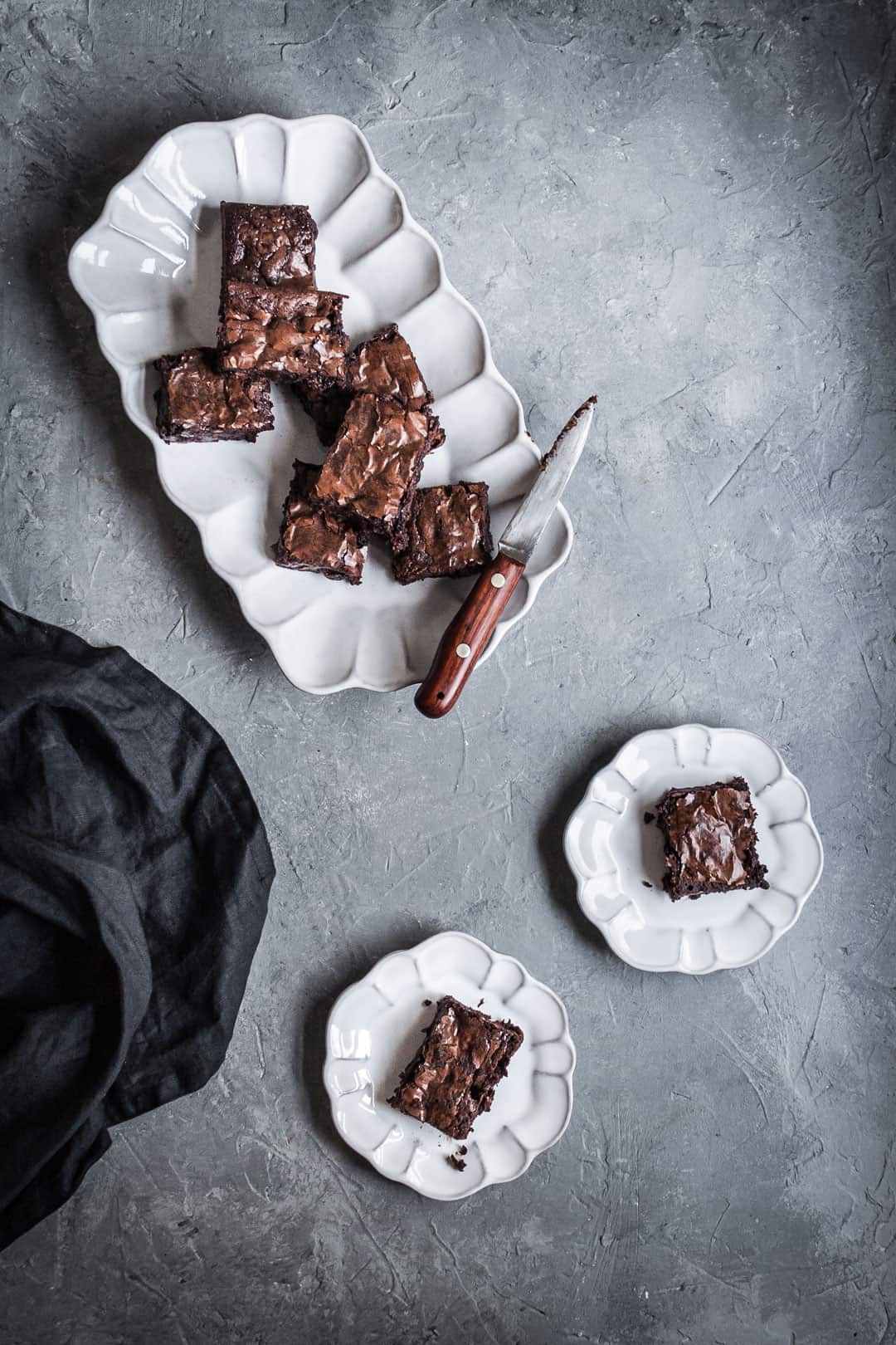 White plates with cut squares of brownies with port soaked cherries, a dark grey linen napkin and a wooden handled knife all on a grey background