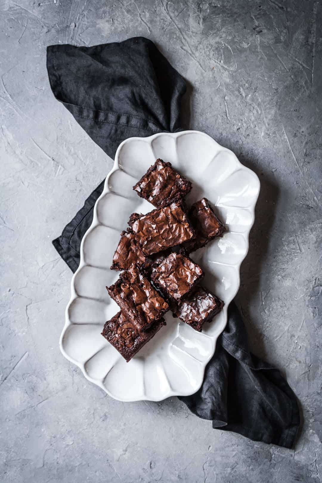 Top view of white fluted platter with cut squares of brownies with port soaked cherries on a grey background with a dark grey linen napkin