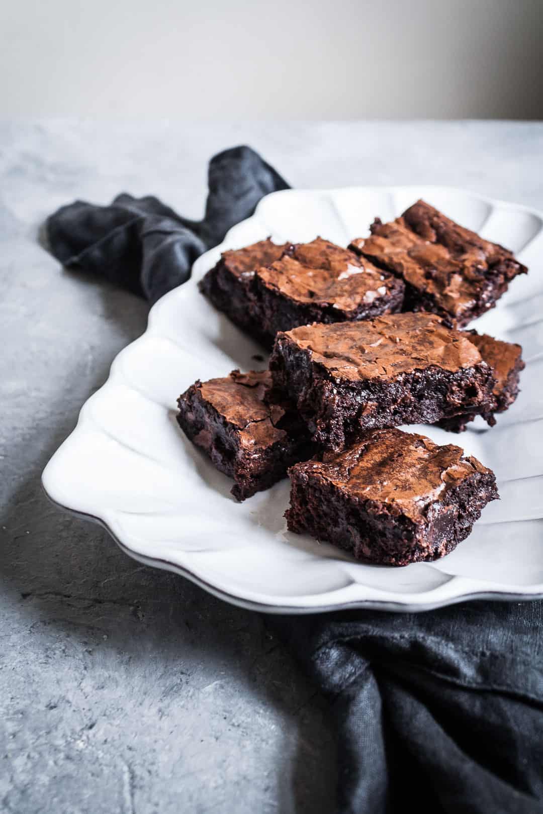 White fluted serving plate with cut squares of brownies with port soaked cherries on a grey background with a dark grey linen napkin under plate