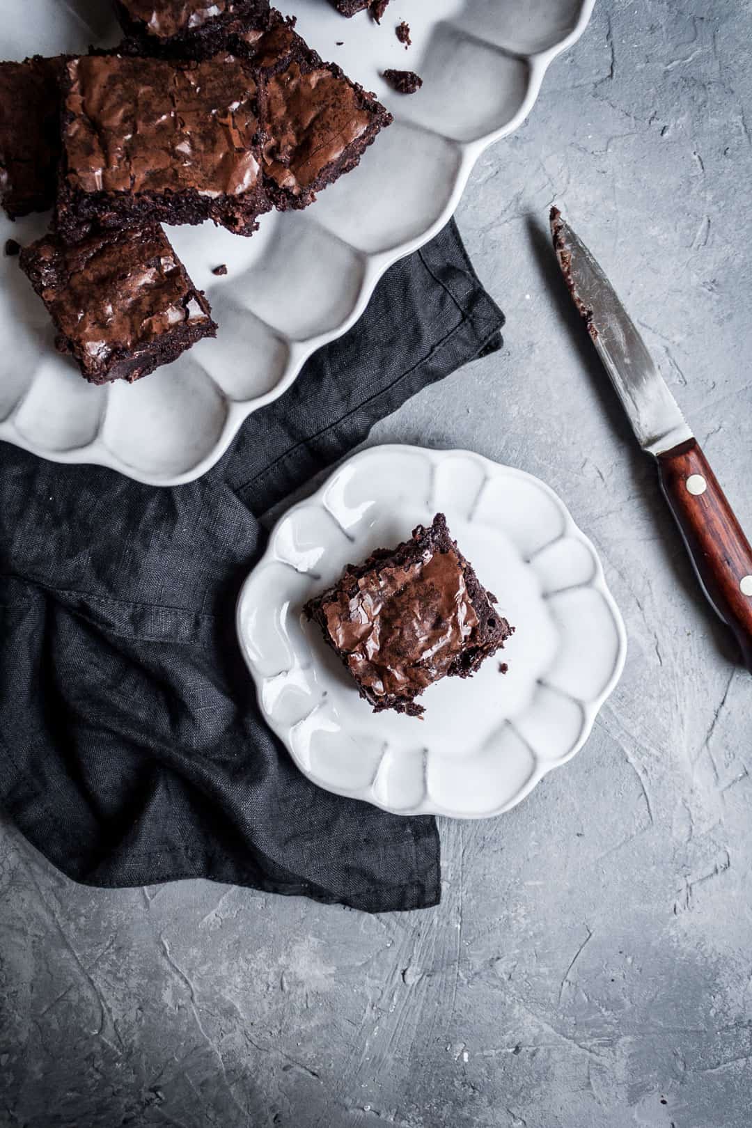 White plate with a cut square of brownies with port soaked cherries, a dark grey linen napkin and a wooden handled knife all on a grey background