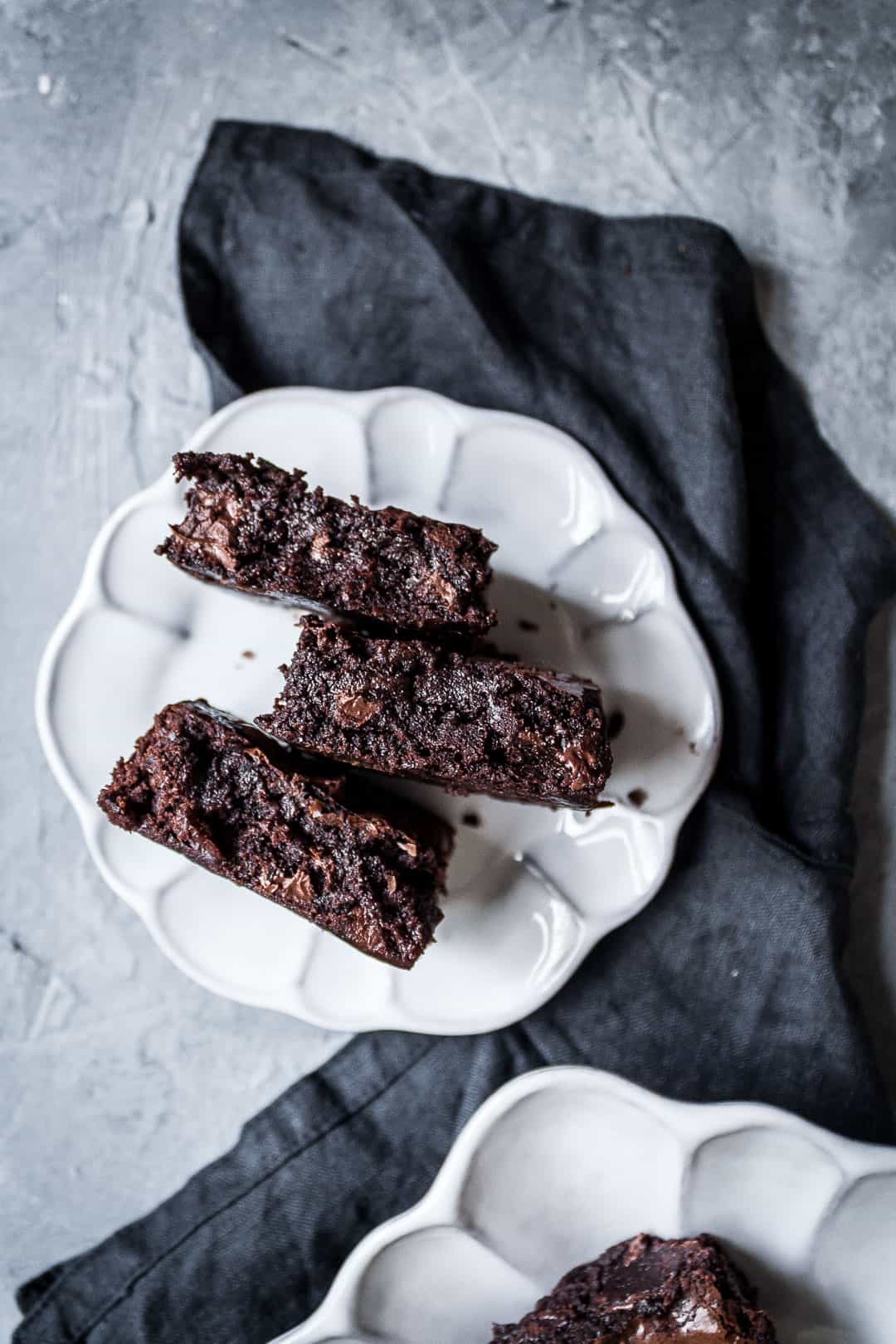 Three cut squares of brownies with port soaked cherries on a white fluted plate with a dark grey linen napkin on a grey background