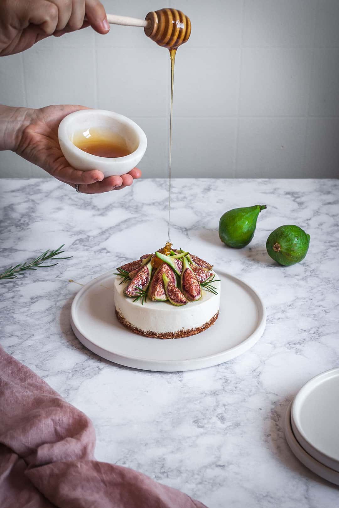 Honey orange cheesecake with figs and rosemary almond crust on a marble backdrop - angled shot of hand holding honey bowl drizzling honey with white plates, green figs