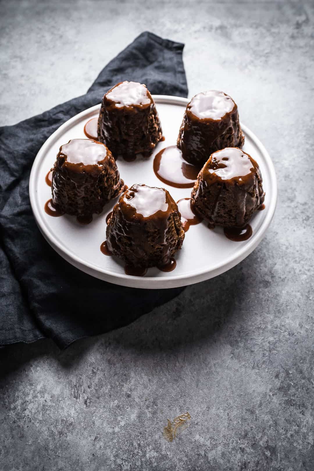 Forty five degree angle view of five backlit apple ginger mini cakes topped with salted caramel sauce on a white plate with a grey background