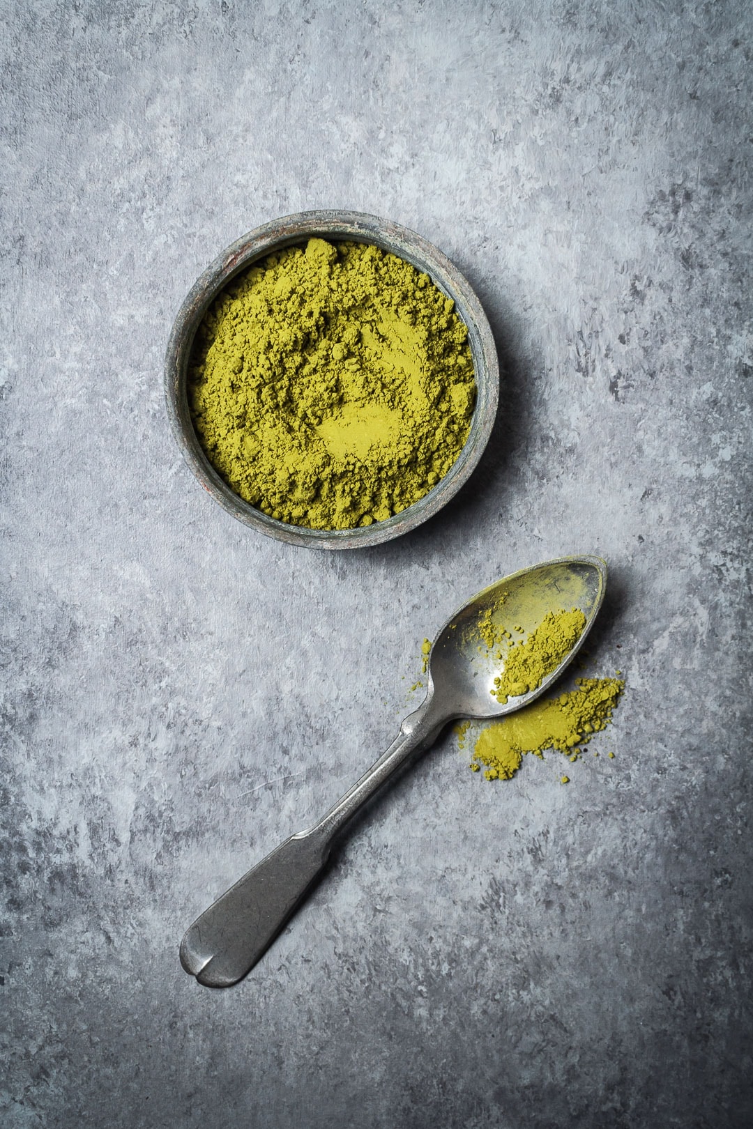Matcha green tea powder in a grey container on a grey background with spoon nearby - recipe for Mint Matcha Chocolate Thumbprint Cookies