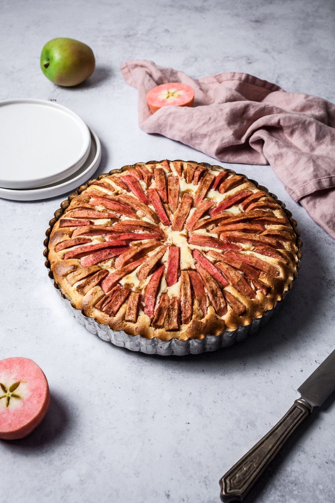 Forty five degree angle view of the Pink Apple Tart with Cream Cheese Filling on a grey background with plates and napkins nearby