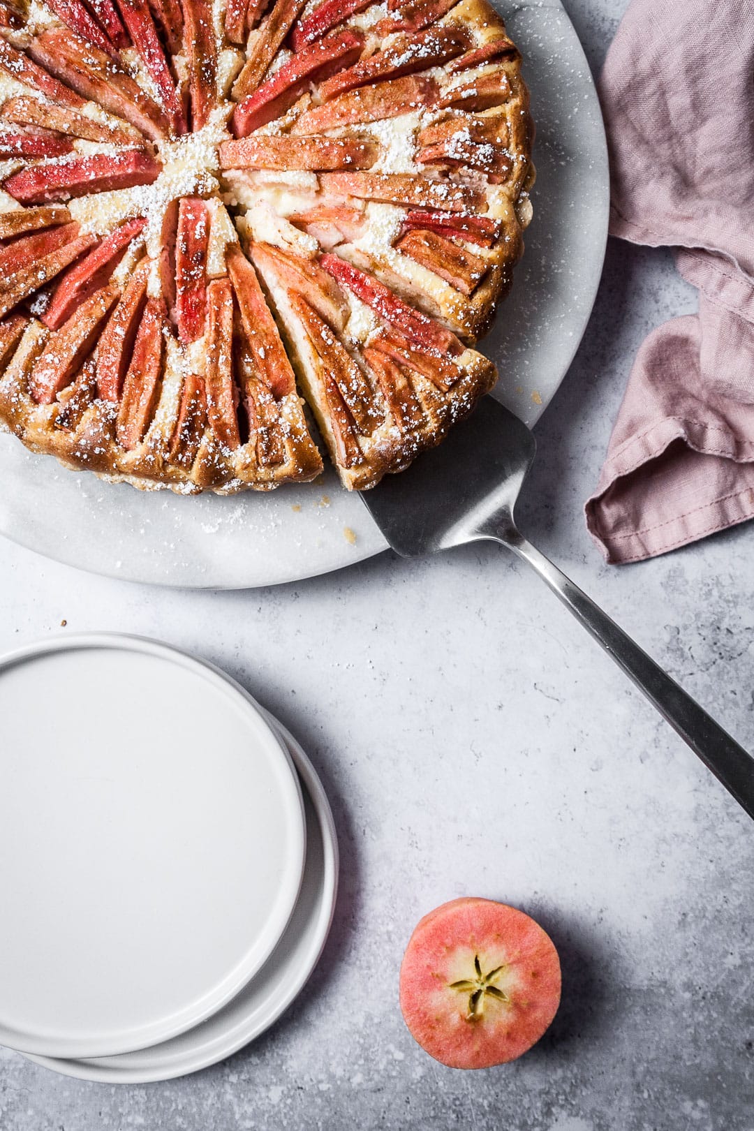 Closeup shot of Pink Apple Tart with Cream Cheese Filling with a slice being lifted off of the marble serving platter; white plates nearby