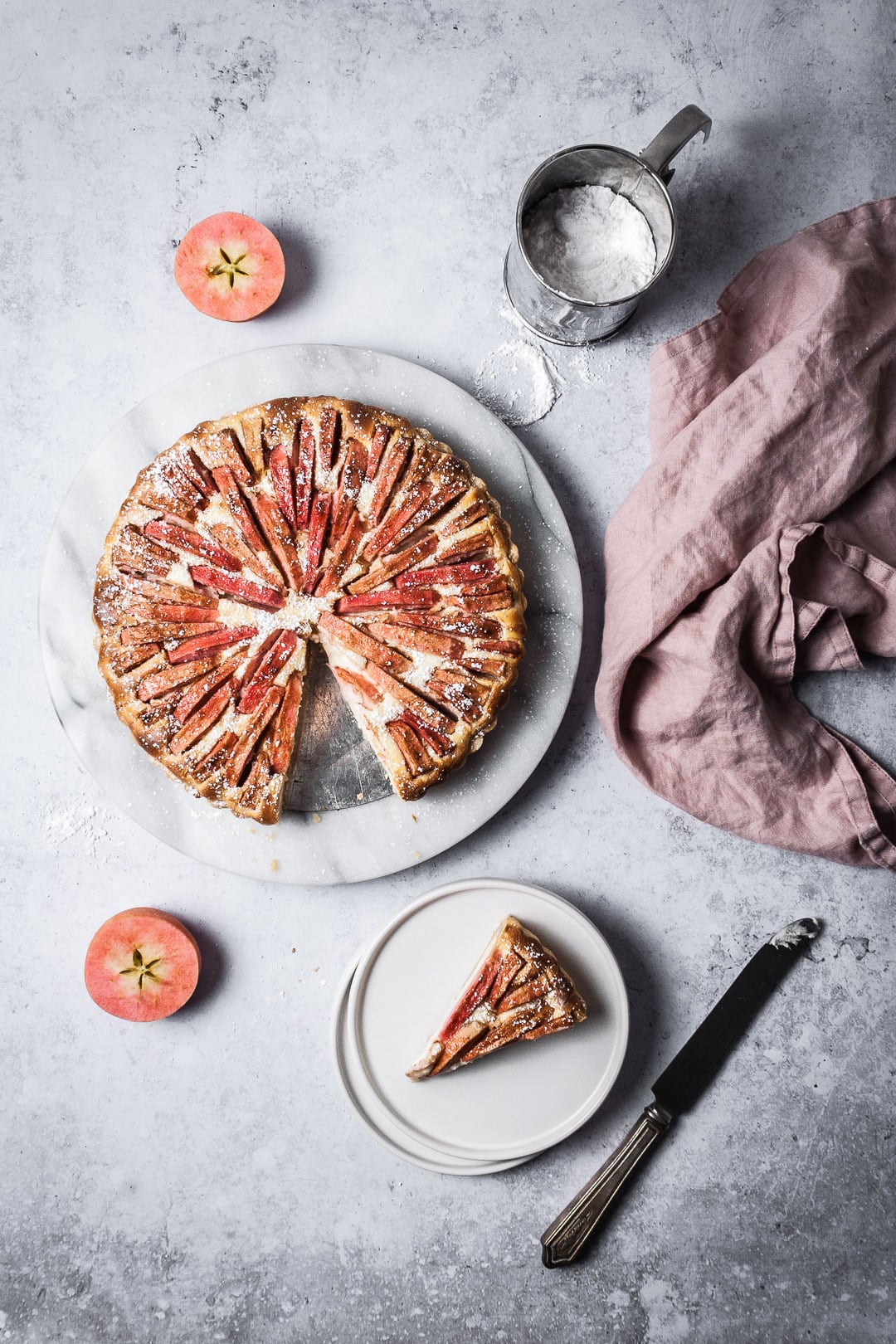 Slice of apple tart on white plates with forks resting nearby; all on a grey background