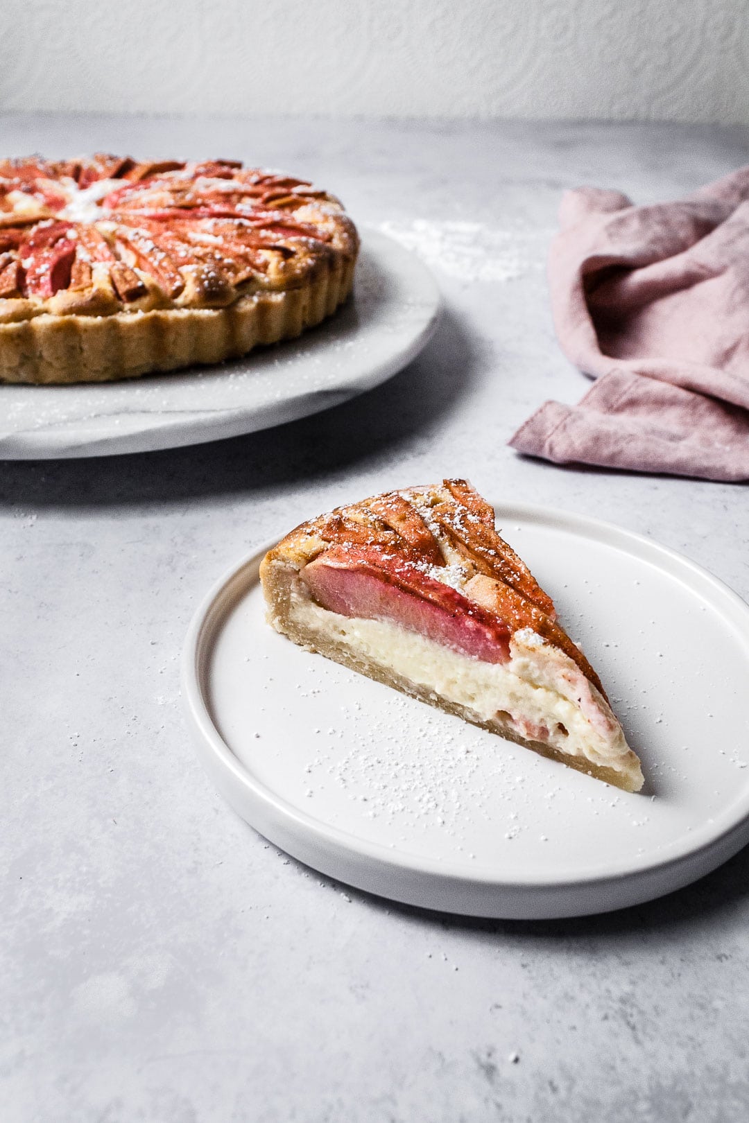 Slice of Pink Apple Tart with Cream Cheese Filling on a white plate and a grey background with whole tart behind it