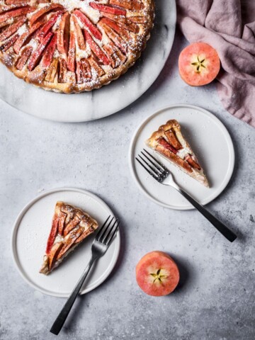 Two slices of Pink Apple Tart with Cream Cheese Filling on white plates with forks resting nearby; all on a grey background