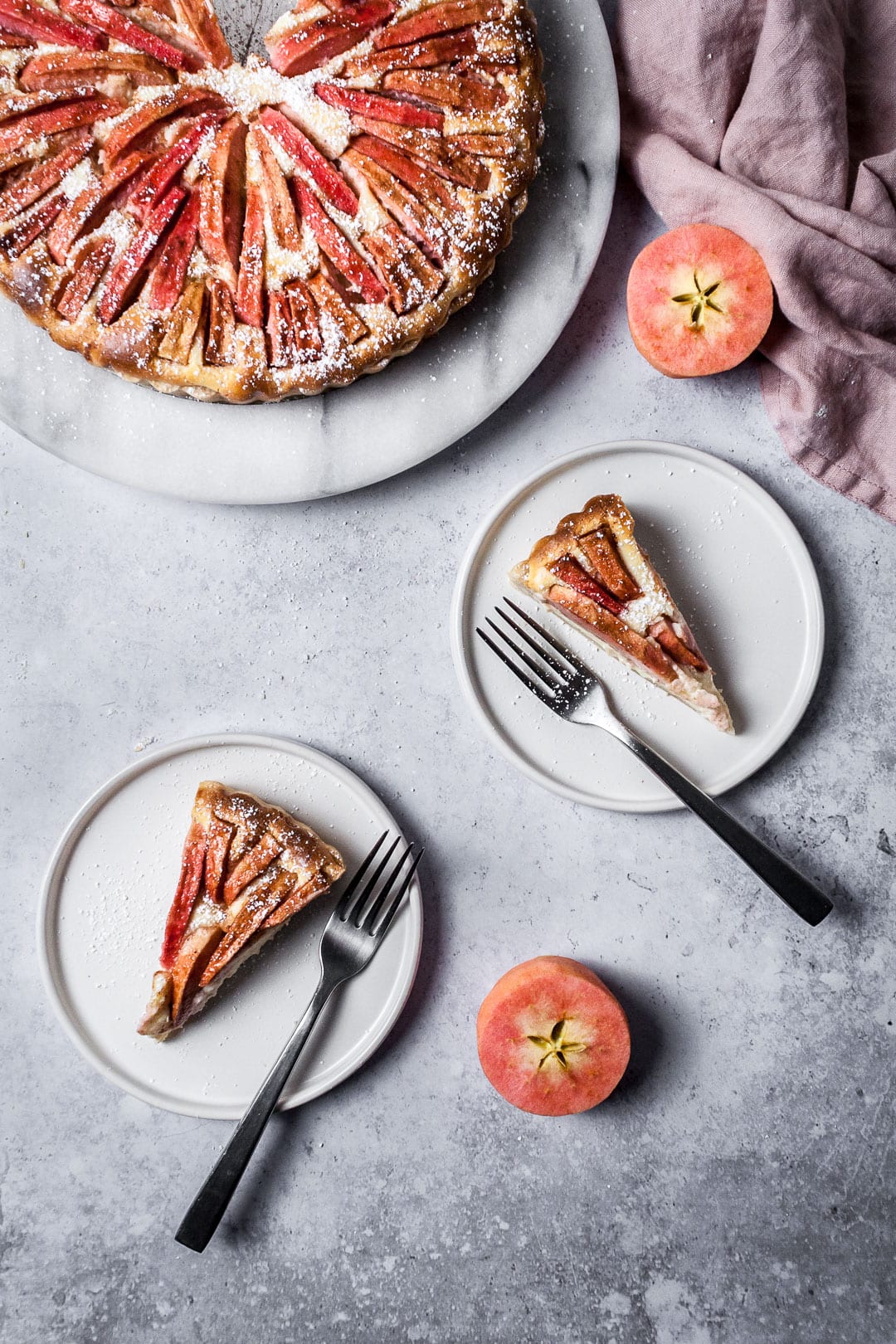 Two slices of Pink Apple Tart with Cream Cheese Filling on white plates with forks resting nearby; all on a grey background