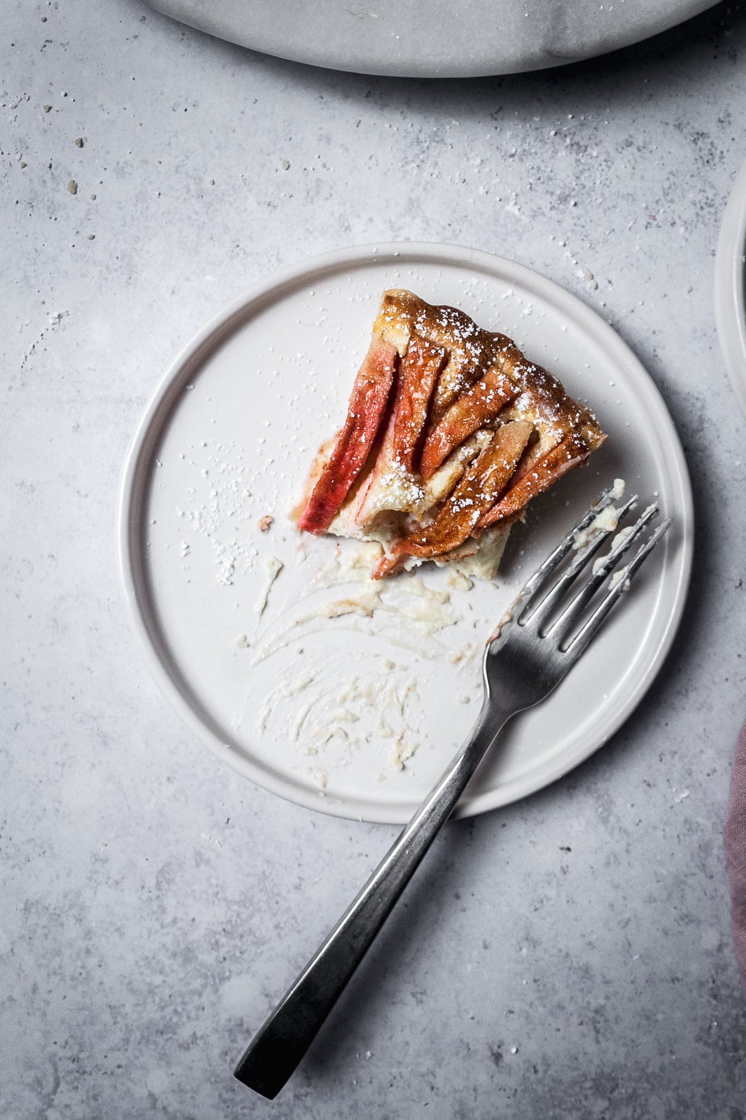 Partially eaten slice of Pink Apple Tart with Cream Cheese Filling on a white plate with a fork, all resting on a grey background