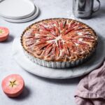 Pink Apple Tart with Cream Cheese Filling on a marble cake stand with a grey background; pink apples cut in half nearby