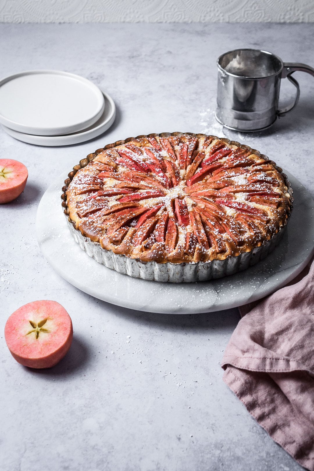 Pink Apple Tart with Cream Cheese Filling on a marble cake stand with a grey background; pink apples cut in half nearby