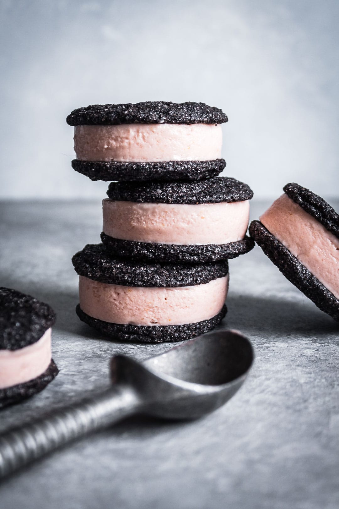 Stack of three ice cream sandwiches with others nearby and ice cream scoop in foreground on grey background